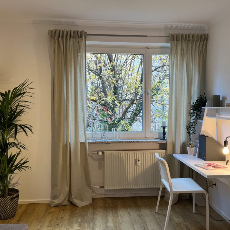 Heller gemütlicher Raum, dekoriert mit Vorhängen, mit einem Schreibtisch, einer großen Pflanze und einem Fenster mit Blick auf Bäume.