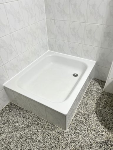 Weiße Duschwanne auf einem grauen Fliesenboden, mit einem Ablauf in der Mitte.