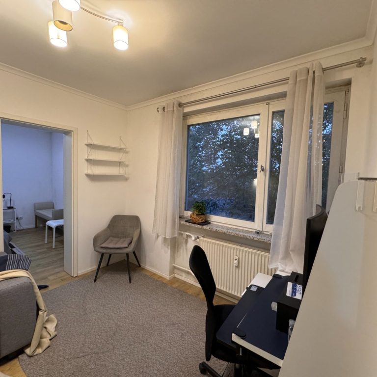 Heller Raum mit Schreibtisch, Stuhl und einem Fenster mit Blick nach draußen.