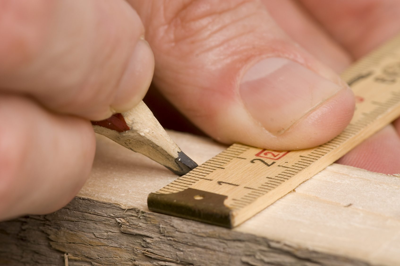 Eine Hand hält einen Bleistift und markiert eine Linie auf einem Holzstück mit einem Zollstock. Maßgetreu genau präzise Arbeiten