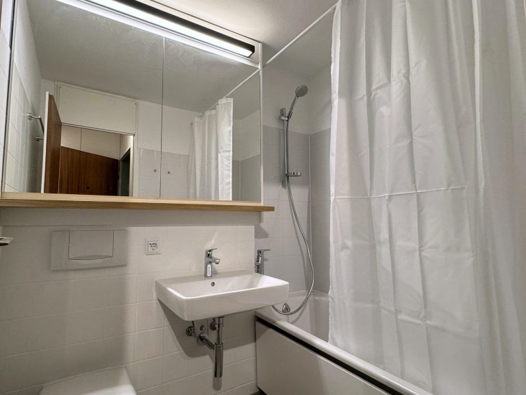 Kleines Badezimmer mit Dusche, Waschtisch und Spiegel mit Holz-Ablage, helle Wände und Vorhang.